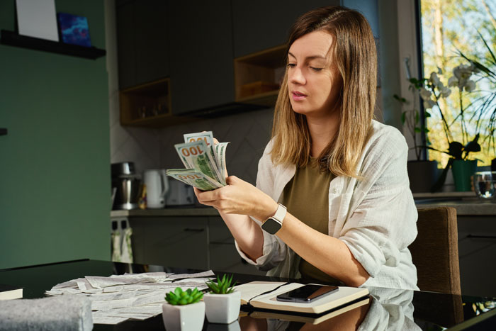 Woman counting money at a kitchen table, surrounded by papers and plants, related to wedding and trip finances.
