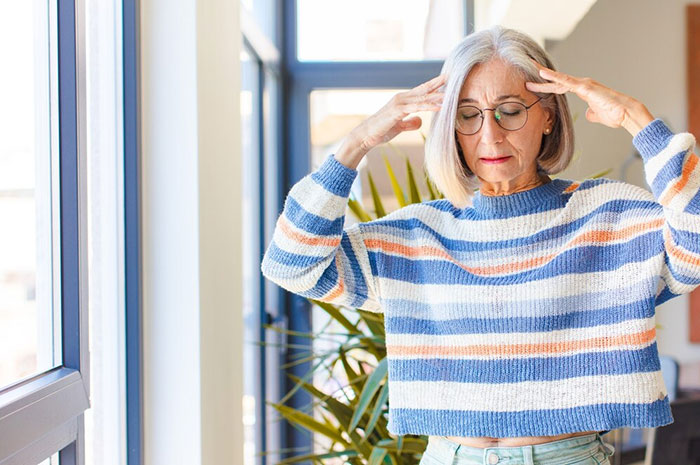 Elderly woman in a striped sweater, standing by a window, practicing a mindful life-saving technique indoors.