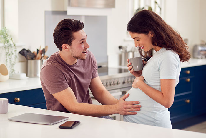 A couple in a kitchen sharing a happy moment, with the man touching the pregnant woman's belly, focusing on pregnancy.