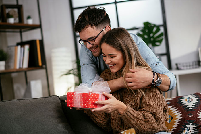 A couple on a sofa, smiling and holding a red gift, highlighting reactions to pregnancy.