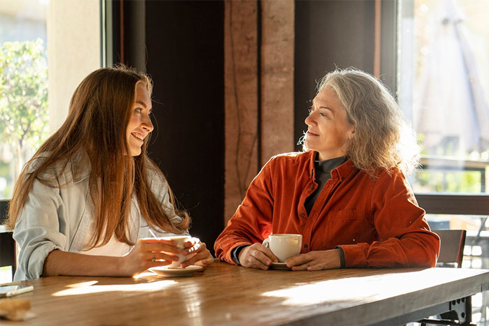 Two women having coffee and chatting in a cozy cafe, with sunlight streaming in through the window.