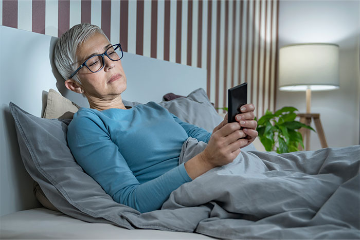 Woman in bed with a smartphone, wearing glasses and a blue top, possibly pondering comments made to pregnant women.