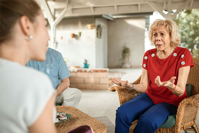Senior woman in red shirt gesturing while talking to a pregnant woman, seated on a porch, discussing pregnancy comments.