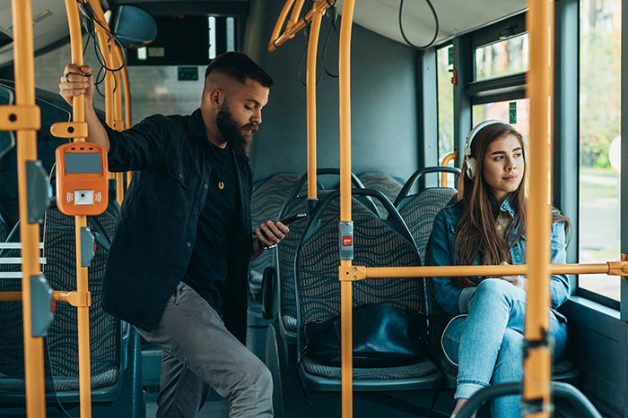 Man and woman on a bus; man holds phone while woman listens to music, illustrating public transport setting.