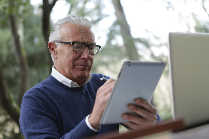 Elderly man with glasses using a tablet outdoors, seated near a laptop.