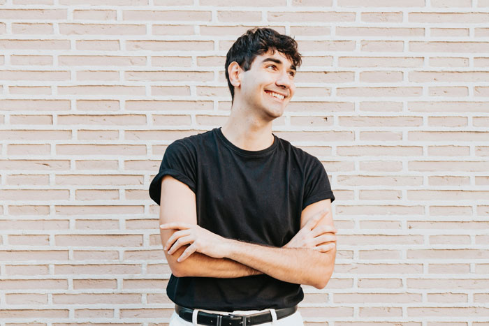A smiling man in a black shirt stands against a brick wall outdoors, illustrating a story on being gay.