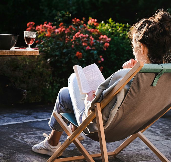Person reading in a deck chair, surrounded by flowers, with a glass of wine nearby, embodying relaxing beauty hacks.