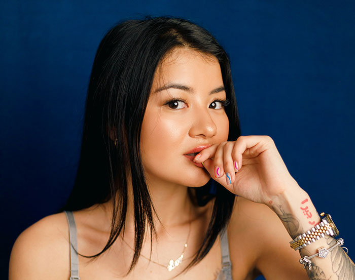 Young woman with tattoos and colorful nails, posing playfully against a blue background.