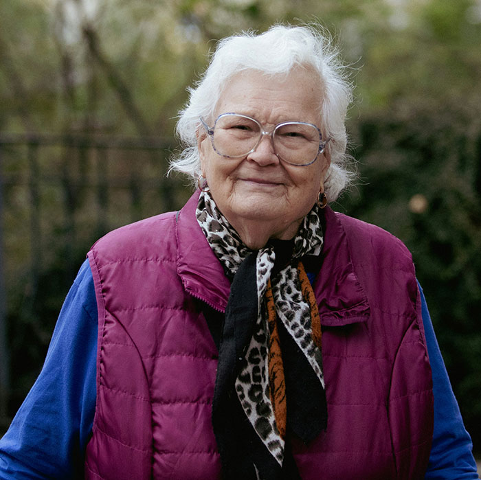 Elderly woman outdoors, wearing a magenta vest and patterned scarf, embodying unique beauty hack style.