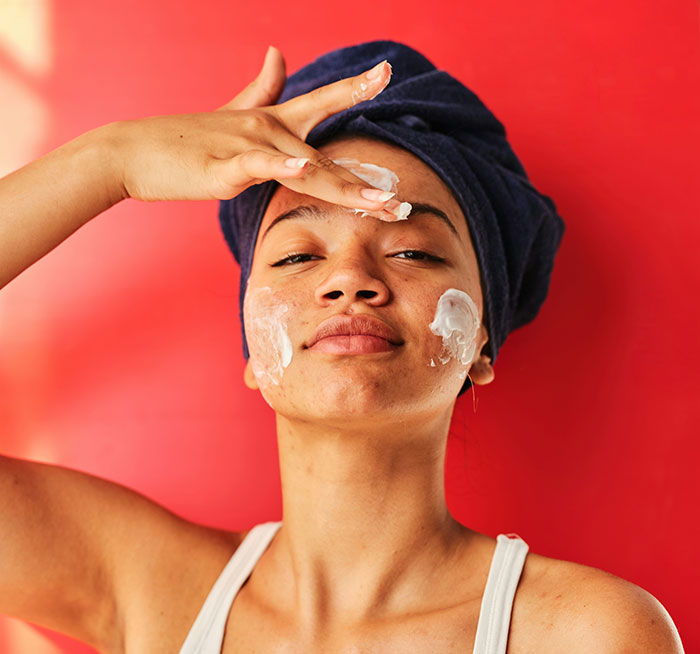 Person applying a beauty hack with cream on face, hair wrapped in towel, against a red background.
