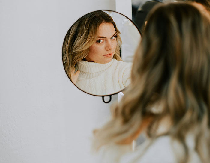 Woman in a white sweater looking into a round mirror, illustrating an unhinged beauty hack exploration.