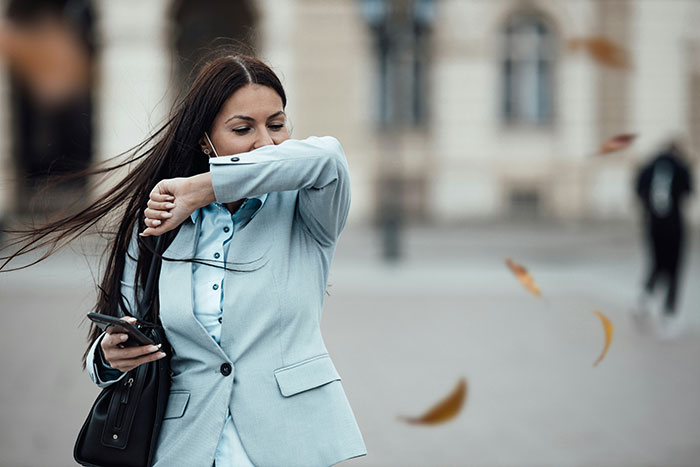 Woman outside sneezing into arm, holding phone; example of unhinged beauty hack against pollution.