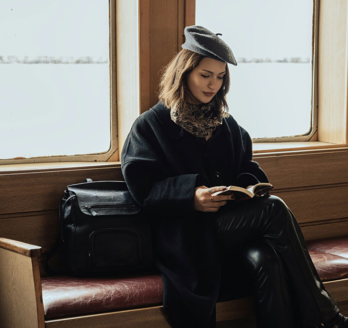 Woman in a black coat and beret reading a book, showcasing unique beauty hacks on a train journey.