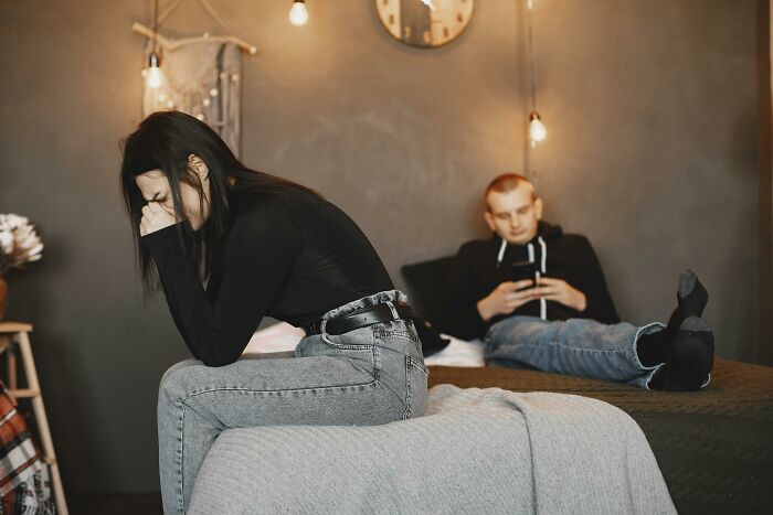 Woman upset and crying on bed while man sits in background on phone, illustrating things exes did that still make blood boil.