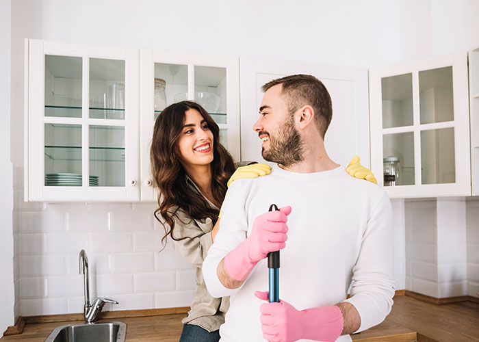 Couple in kitchen smiling, wearing gloves for cleaning, representing wild marriage hacks for household chores.