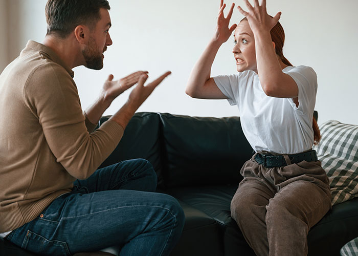Couple on a couch, animatedly discussing marriage hacks, displaying frustration and gestures.