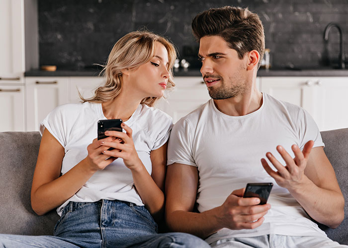 Couple on a couch using phones, exchanging curious looks, illustrating unique marriage hacks dynamics.