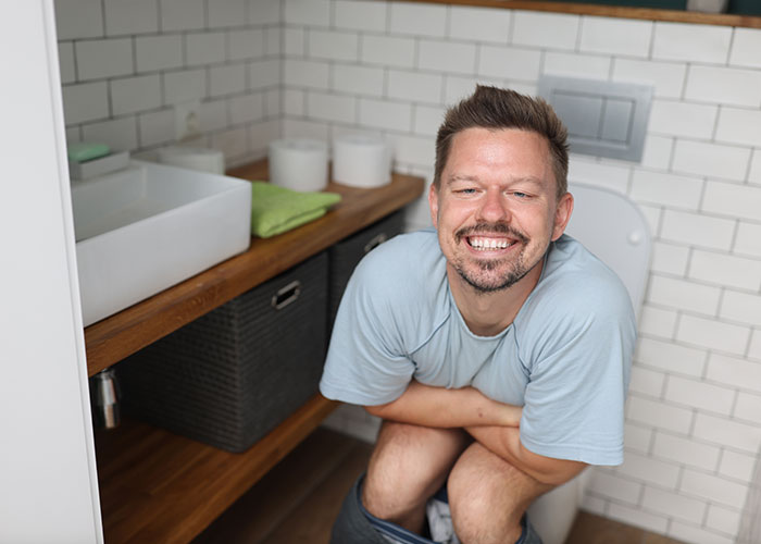 Man smiling while sitting on toilet, showcasing unique marriage hacks in a small bathroom setting.