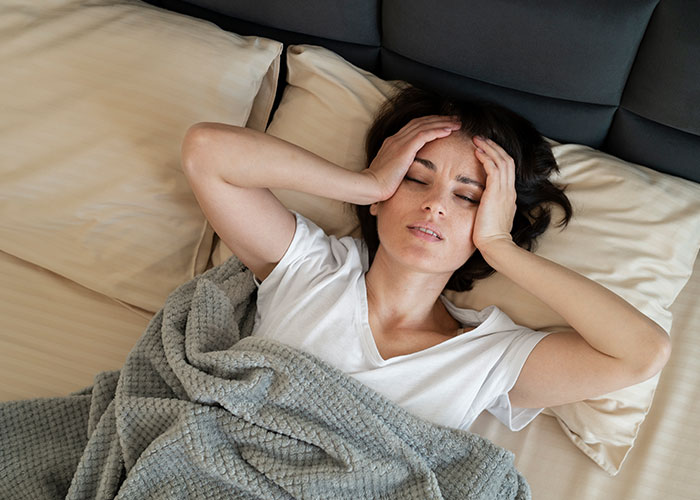 Woman in bed with a headache, using a unique marriage hack for relaxation under a gray blanket.