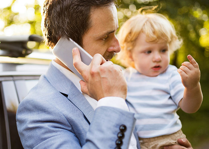 Man holding a child while talking on the phone outdoors, showcasing ways women "decenter" men in everyday life.