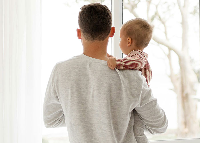 Man holding a baby by a window, wearing a light sweater, symbolizing women who "decenter" men in family life.