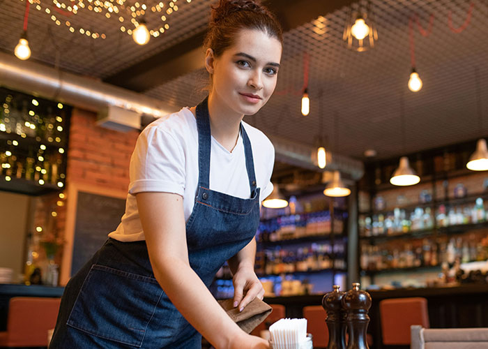 A woman in a restaurant wearing an apron, confidently setting a table, illustrating how women decenter men.
