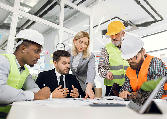 Group of professionals in a meeting, with a woman leading the discussion, wearing construction helmets.