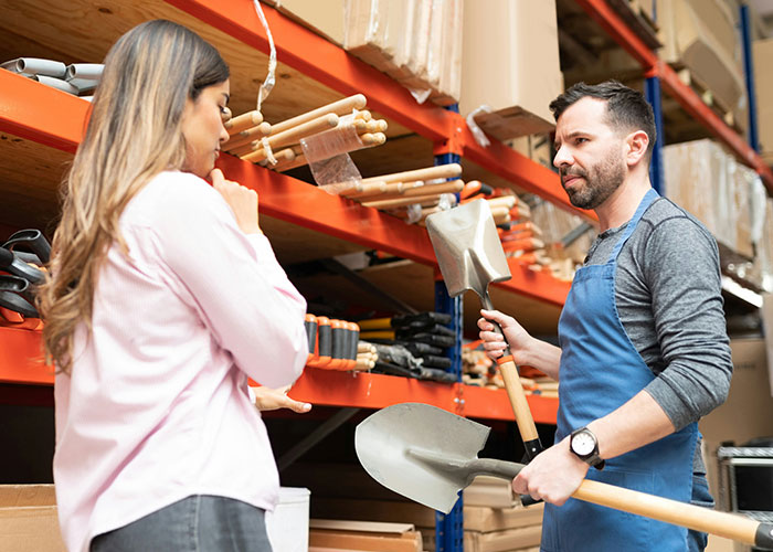 Woman observing a man holding shovels in a warehouse, highlighting ways women decenter men's focus.