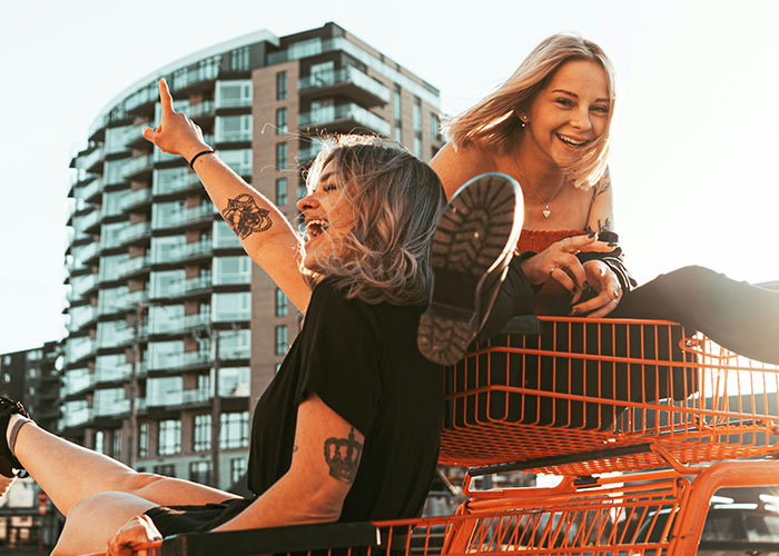 Women having fun in a shopping cart, emphasizing how they decenter men, with a modern building in the background.