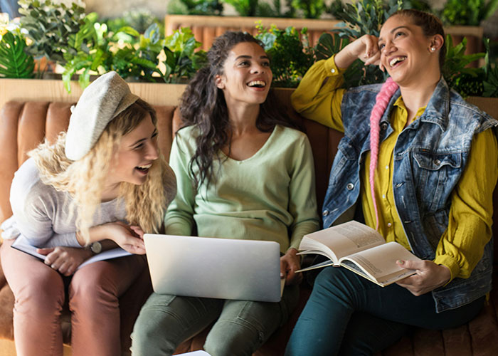 Women laughing together, using a laptop and book, in a cozy setting, illustrating ways to decenter men.