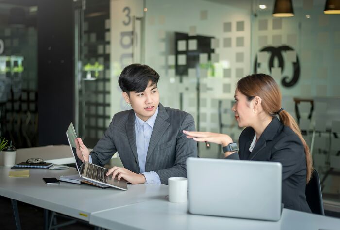 Two coworkers in suits discussing at a table, with laptops and coffee cups, in a modern office setting.