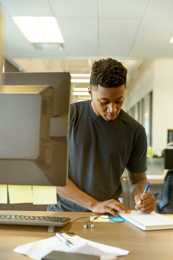 Person writing in notebook at an office desk, dealing with coworkers.