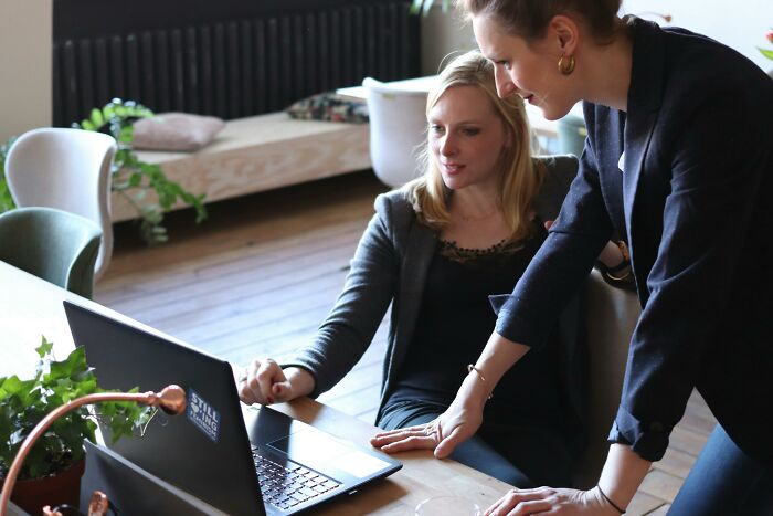 Two coworkers in an office setting, one pointing at a laptop screen, demonstrating workplace interaction dynamics.