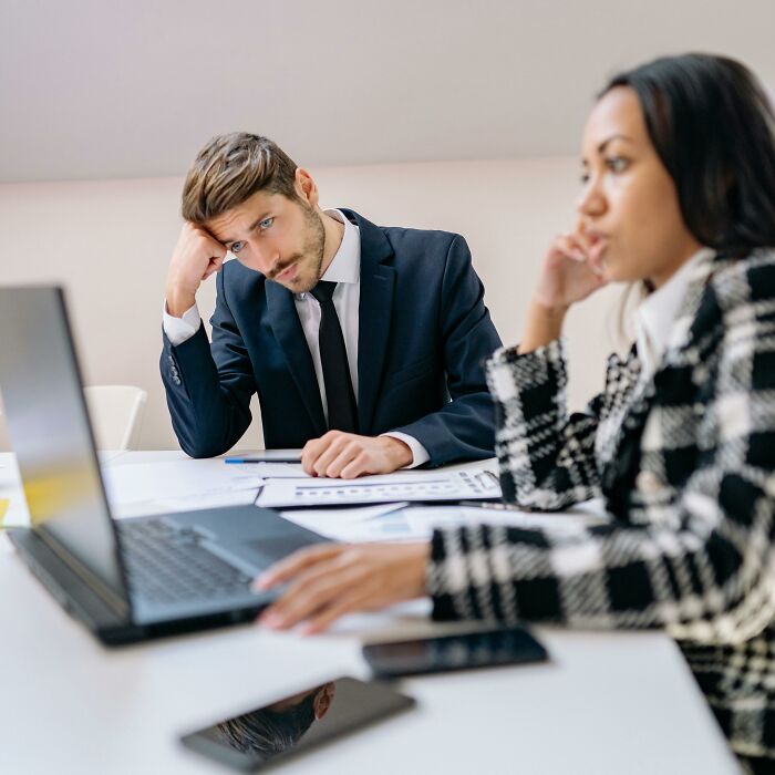 Man in suit looking frustrated at work, colleagues discussing ways to deal with coworkers.