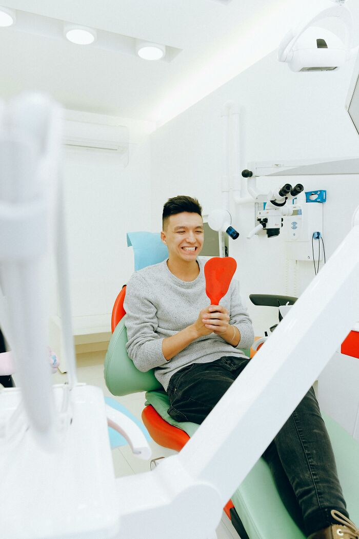 Person smiling in a dentist chair, holding a red mirror, in a modern clinic.