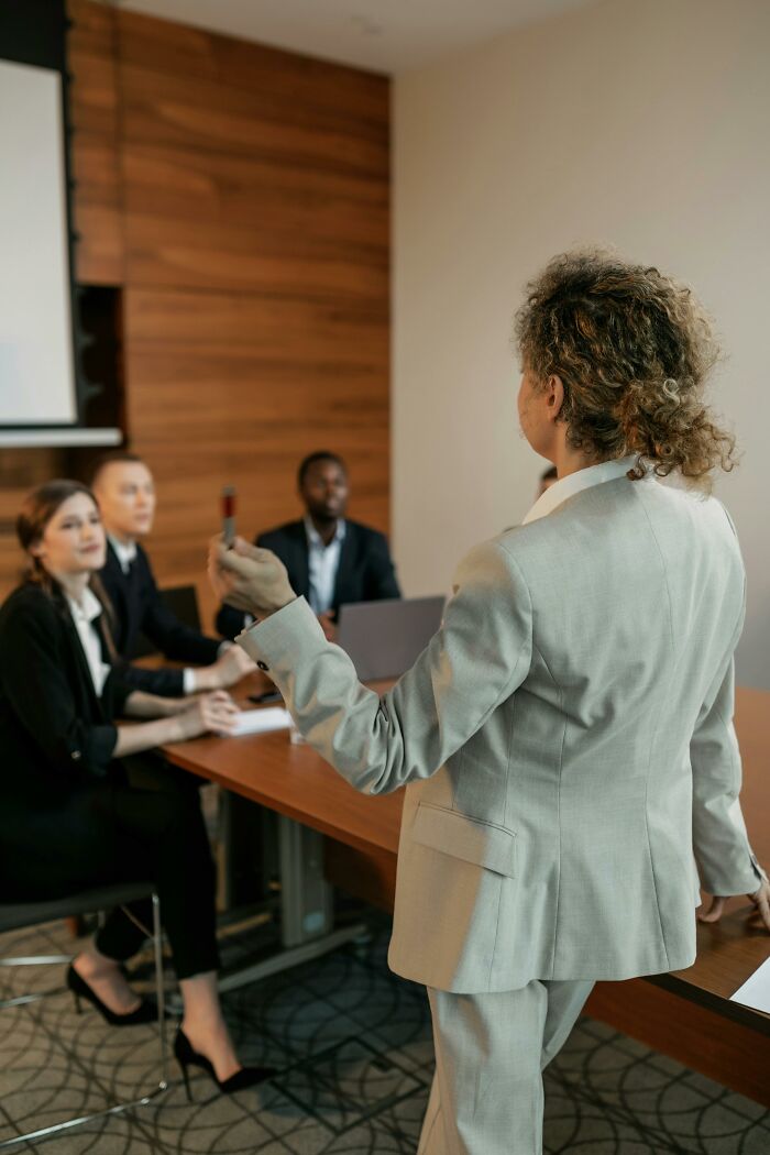 A person in a suit presenting at a table, discussing expensive weddings with attentive listeners.