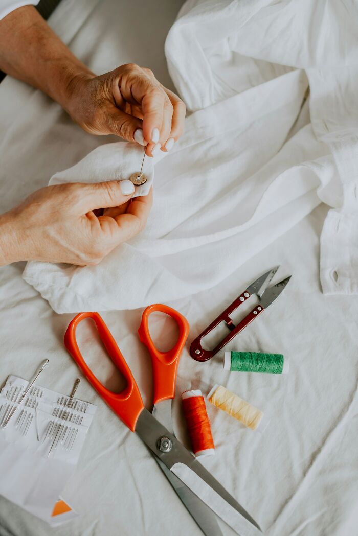 Hands sewing a button on a shirt surrounded by scissors and thread, symbolizing expensive weddings' DIY projects.