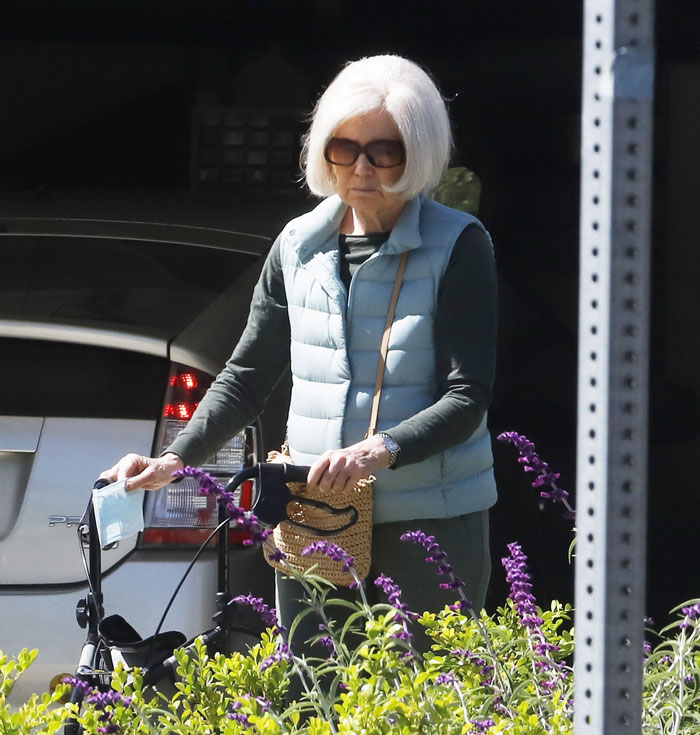 Mary Kay Place casually strolls with a walker, wearing sunglasses and a light blue vest, surrounded by purple flowers.