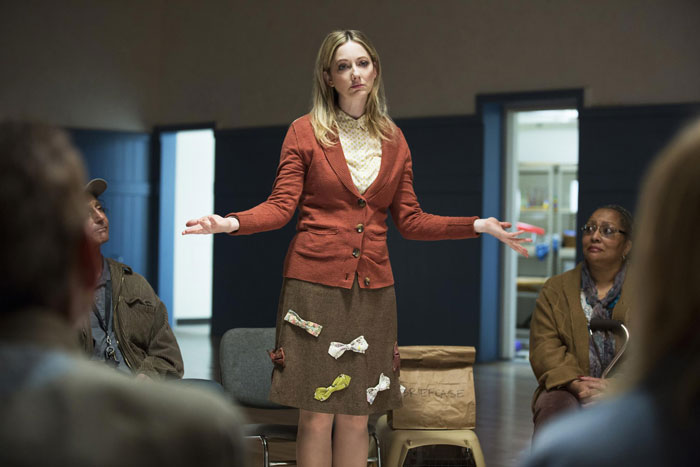 Woman in a classroom setting, wearing a brown and red outfit, gestures with arms outstretched.