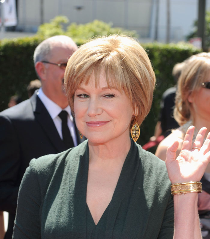 Mary Kay Place smiling outdoors in a green outfit, waving to the camera.