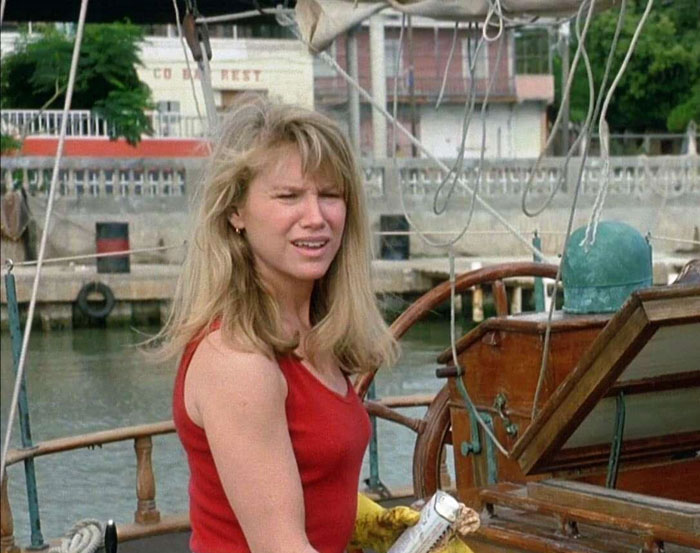 A woman with blonde hair in a red tank top, standing on a boat, wooden deck visible in the background.