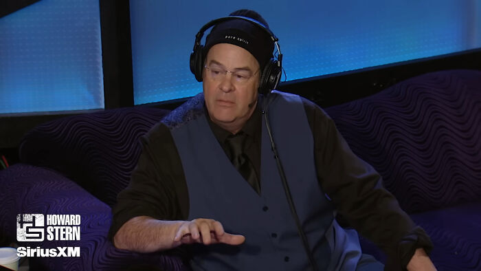 Man at a radio show, wearing headphones and a blue vest, speaking on air.