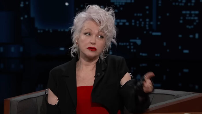 A celebrity with white hair in a TV interview, wearing a black jacket and red top.