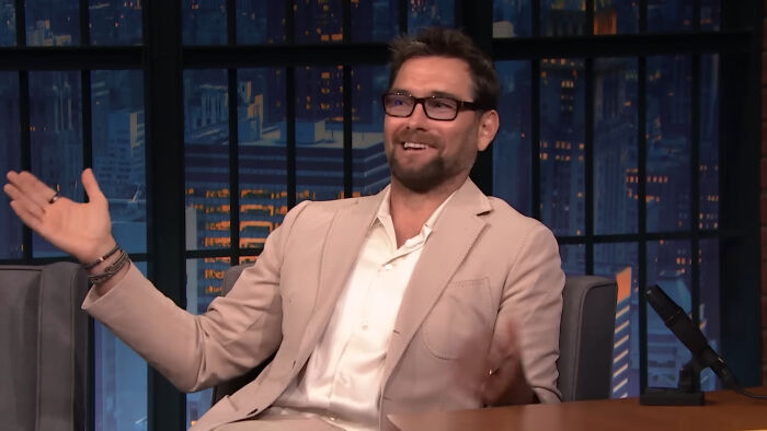 A celebrity in a beige suit and glasses smiling on a talk show set.