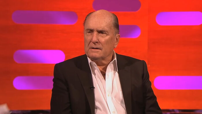 A celebrity sitting on a talk show set, discussing with the host against a vibrant red and purple background.