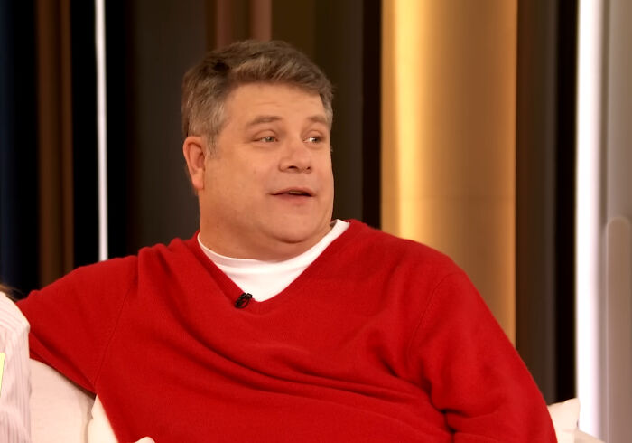 A celebrity in a red sweater sits on a couch, engaging in a casual conversation.