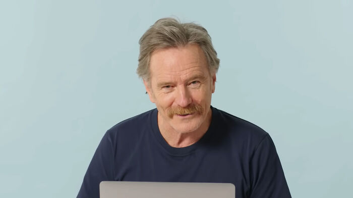 Smiling celebrity with gray hair and a mustache against a light blue background, showcasing a friendly demeanor.