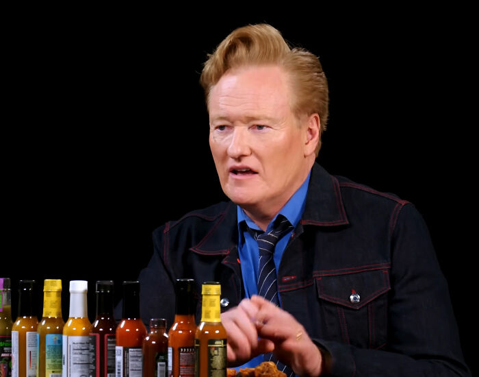 Celebrity enjoying hot sauces during an interview, wearing a blue shirt and dark jacket.