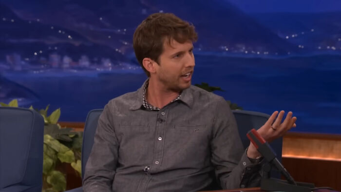A celebrity talking animatedly on a talk show set, wearing a gray shirt.