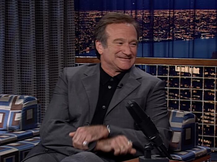 Smiling celebrity in a suit during a talk show appearance, seated and engaging warmly with the audience.